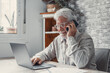 © Daniel - Portrait of smiling mature man sit at table at home talk on cellphone work on computer gadget. Smart senior 70s male have smartphone call conversation, use laptop. Elderly and technology concept..