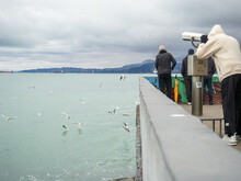 Man On The Pier Against The Backdrop Of Seagulls. People Feed The Seagulls.  Beautiful Seascape In Cloudy Weather