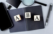 Word ABA on wooden block on black notebook with smartpone, credit card and magnifier