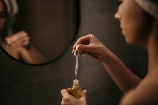 young woman looking in the mirror, holding a dropper with hyaluronic acid serum close to her face. s