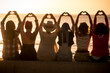 © simona - Group of women sitting against a golden sunset and doing heart symbol with hands and raised arms for friendship and love freedom concept. Women international day. Travel and vacation. Friends at beach
