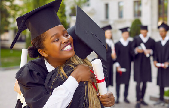 Congrats, grad. Proud of you. Happy, joyful university graduates celebrate graduation together. Beautiful Afro American student girl in square black cap holding scroll, hugging her friend and smiling