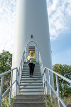 Female Engineer With Brown Curly Hair, Yellow Helmet And Digital Tablet Looking At Camera While Walking Down The Stairs Of A Modern Renewable Energy Wind Turbine After Inspection