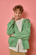 © Folenial - Vertical studio shot of relaxed cute freckled man with curly red hair smiles happily keeps hands near face thinks about something pleasant wears casual green zipped hoodie isolated on pink background.