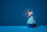 Fototapeta Tęcza - Blue cupcake with red and white sprinkles and lit sparkler on a blue background.