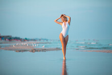 Full-length Portrait Of A Beautiful Sexy Young Slim Caucasian Blonde Girl In White Swimsuit Standing In Sea Water With Reflection At The Beach In Warm Sunset Light With Softly Blurred Background