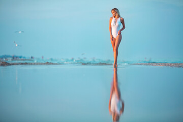 Wall Mural - Full-length portrait of a beautiful sexy young slim caucasian blonde girl in white swimsuit standing in sea water with reflection at the beach in warm sunset light with softly blurred background