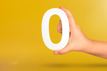 Fototapete - Numeral zero in hand. A hand holds a white number zero on a yellow background with copy space. Zero concept, 0 percent interest rate, minimum air emissions, cost or credit no increase.