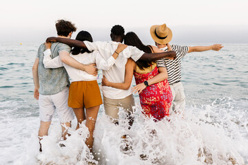 View from back of group of young diverse people friends hugging each other and enjoying sunset at beach in summer holidays