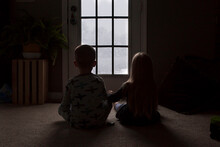 Rear View Of Siblings Looking Through Window While Sitting On Floor In Darkroom
