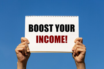 Boost your income text on notebook paper held by 2 hands with isolated blue sky background. This message can be used as business concept about increasing your income.