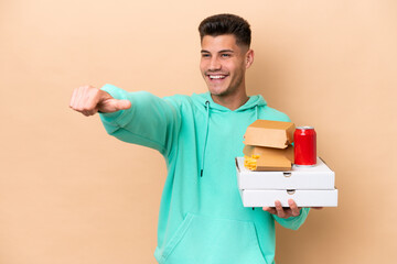 Wall Mural - Young caucasian man holding fast food isolated on beige background giving a thumbs up gesture