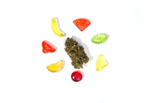 A Dry Bud Of Marijuana Lies On A White Background Surrounded By Chewy Gummy Candies Of Various Shapes And Colors On A White Background.  Top View, Lots Of Empty Space