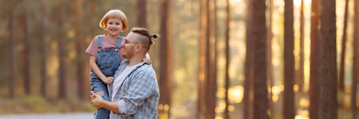 Dad holds a little daughter in his arms, they laugh merrily walking along a forest road among tall pines. Family walk in the forest at sunset. Banner for website header design with copy space.