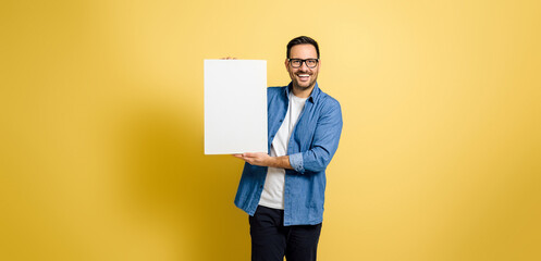 Mid adult cheerful businessman holding blank white banner template mock up and smiling at the camera while standing isolated against yellow background