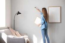 Young Woman With Blank Frame Nailing On Grey Wall At Home