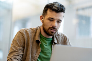 Pensive, attractive man using laptop computer, having video conference. Student in wireless earphones studying, learning, sitting in library, online education