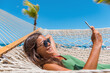 © Maridav - Happy beach woman using phone app reading sms texting lying on hammock relaxing on tropical vacation. Young casual cute girl lying down in outdoor swing bed enjoying sun sunbathing using mobile phone