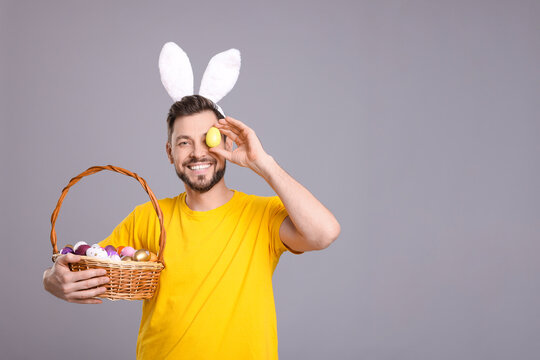 Wall Mural - Happy man in bunny ears headband holding wicker basket with painted Easter eggs on grey background. Space for text