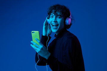 Wall Mural - Man smile with phone in hand taking selfies with headphones listening to music, portrait dark blue background, neon light, style and trends, mixed light, men's fashion, copy spot
