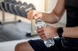 Sport man with well trained body in black sportswear opening water bottle while sitting on workout bench in fitness gym. Drinking water after weight training exercise.