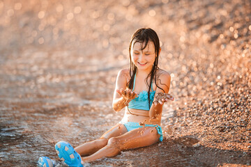 Little happy girl playing with sand on the seashore. Smiling child is sunbathing while sitting by the water.