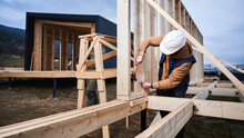Man Worker Building Wooden Frame House On Pile Foundation. Carpenter Hammering Nail Into Wooden Truss, Using Hammer. Carpentry Concept.