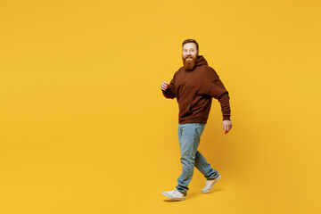 Wall Mural - Full body smiling happy fun cheerful satisfied young redhead caucasian man wearing brown hoody casual clothes walk go look camera isolated on plain yellow background studio portrait Lifestyle concept