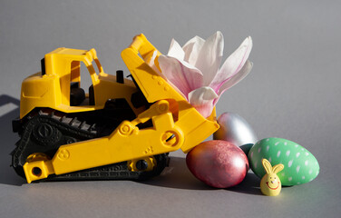 Wall Mural - toy orange bulldozer, magnolia flower, colorful easter eggs, easter bunny on a gray background. Happy Easter concept for business, construction companies soft focus. spring revival