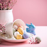 Fototapeta Zwierzęta - White porcelain coffee cup with colorful quail eggs and spring flowers over pink background. Springtime and Easter holiday concept with copy space