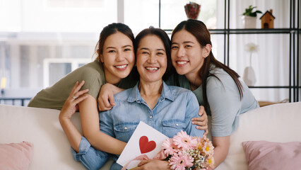 Happy time Mother day grown up child looking at camera cuddle hug give flower gift box red heart card to mature mum. Love kiss care mom asia middle age adult people smile enjoy sitting at home sofa.