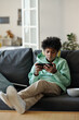 © pressmaster - African American schoolboy in casualawear watching online movie or video in smartphone while having rest on comfortable couch at home
