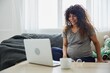 © SHOTPRIME STUDIO - Pregnant woman smile sits at home on the sofa with a laptop and talks on video chat, blogger, freelancer works online. Home clothes, lifestyle of a pregnant woman, preparation for childbirth