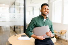 Carefree Inspired Indian Male Employee Standing In Modern Office Space And Holding Laptop, Cheerful Young Businessman In Green Shirt Looks Aside With Light Friendly Smile, Full Of Plans And Ideas