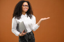 Beautiful African American Business Woman Carrying Laptop And Pointing With Hand Aside, Holding Empty Space Above Hand Palm Standing Isolated On Brown, Female Office Employee Presenting, Advertising