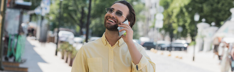 Carefree man in sunglasses talking on cellphone on blurred urban street in summer, banner.