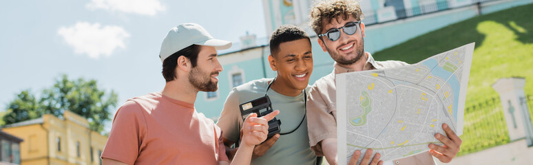 Wall Mural - tourist in sun cap pointing at city map near smiling tour guide and african american man on andrews 