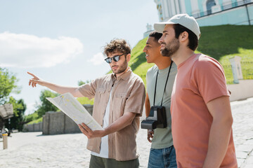 Wall Mural - tour guide in sunglasses holding city map and pointing with finger near multiethnic travelers on and