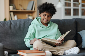 Clever boy with open book sitting on grey comfortable couch in living room and reading interesting novel or story while preparing homework