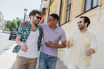 Wall Mural - cheerful multiethnic friends in sunglasses walking on blurred andrews descent in kyiv.