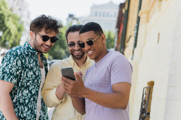 Wall Mural - Multiethnic men in sunglasses using smartphone on urban street in Kyiv.