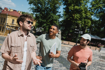 Wall Mural - multiethnic travelers with map and vintage camera listening to young guide while walking on andrews 