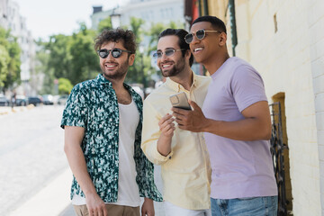 Wall Mural - smiling interracial friends in sunglasses holding cellphone and looking away on street in kyiv.