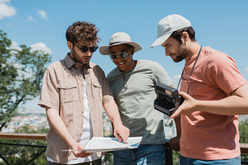 Wall Mural - young tour guide in sunglasses looking at travel map near interracial tourists with vintage camera i