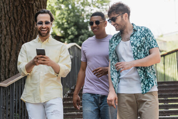 Wall Mural - Cheerful man using smartphone while walking near multiethnic friends on stairs in summer park.