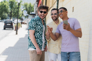 Wall Mural - multiethnic friends in sunglasses looking away and holding smartphone on street in kyiv.