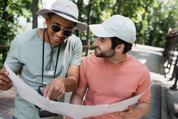 Wall Mural - smiling african american tourist in sunglasses pointing at map near bearded friend in summer park.