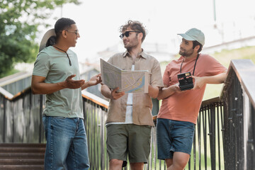 Wall Mural - smiling african american man gesturing while talking to friend holding travel map on stairs of urban