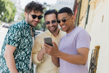 Wall Mural - positive interracial friends in sunglasses using mobile phone on urban street in summer.