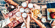 Multi racial friends toasting beer glasses on wooden table covered with food - Top view of people having dinner party at bar restaurant in happy hour- Food and beverage lifestyle concept